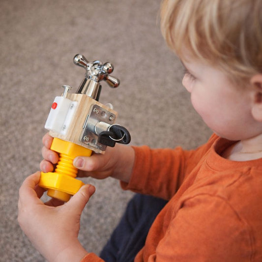 Toddler playing with a Montessori busy toy featuring a turning tap handle and twist mechanisms for fine motor skill development.